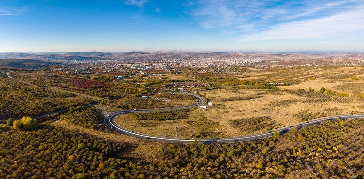 Aerial view of a winding road cutting through the rolling hills with autumn foliage and a distant cityscape in Feleacu, Cluj County, Romania.