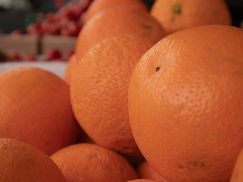 Oranges are stacked together at a market. The sun shines down, making the oranges bright. People walk by, stopping to shop for fruit