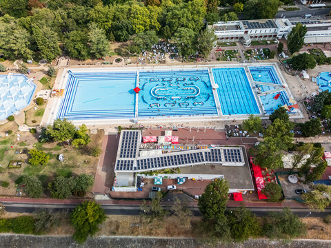 Aerial view of Palatinus lido swimming pools and waterslides on Margaret Island, Budapest, Hungary, with swimmers, sun loungers, pool buildings and surrounding late-summer trees.
