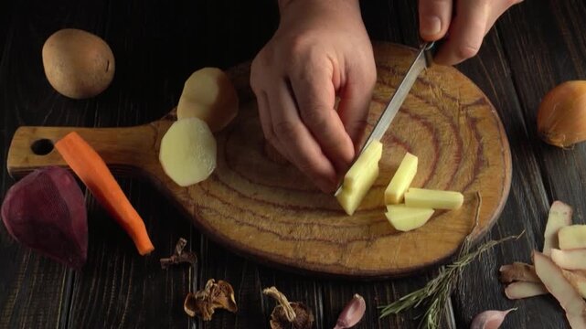 Hands slicing a peeled potato into pieces on a rustic wooden cutting board surrounded by assorted vegetables, herbs, and spices in an overhead kitchen preparation scene
