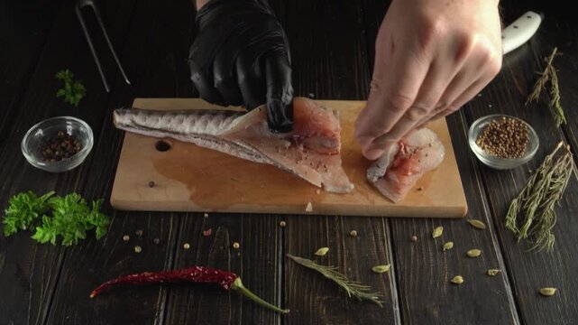 Gloved hands slice raw fish fillet with a kitchen knife on a wooden cutting board surrounded by herbs, spices, and peppers in a rustic overhead food preparation setting