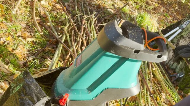 A person inserts a branch into a wood shredder on grassy yard ground. The machine whirs loudly, producing a harsh grinding and crunching sound.