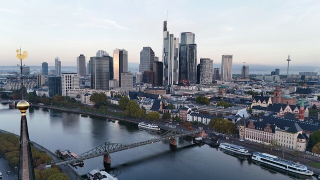 Aerial view of the Frankfurt skyline featuring modern skyscrapers, the Main river, and the Eiserner Steg bridge under a soft morning light in Frankfurt am Main, Hessen, Germany.