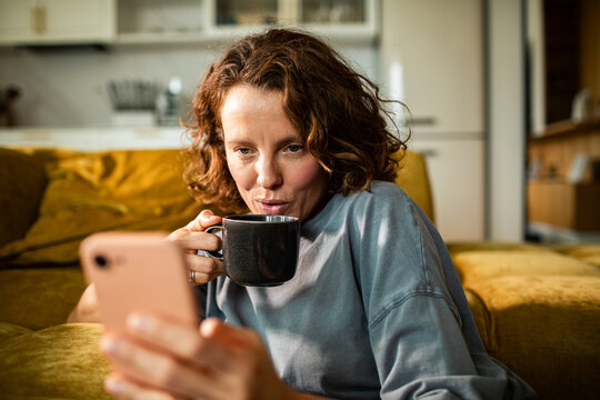 Woman blows on hot coffee while checking phone at home