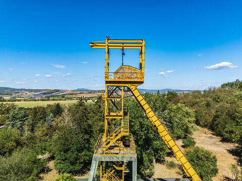 Aerial view of the historic Tiribesi-Akna coal mine headframe rising above trees and rolling farmland in Batonyterenye, Hungary, during summer.