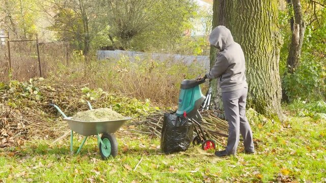A person feeds branch into a garden shredder in a grassy backyard. A wheelbarrow stands nearby filled with wood chips. The machine makes a loud grinding noise.
