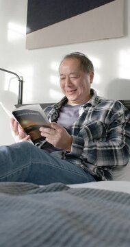 Vertical video: Senior Asian man sitting on bed sun casting stripes reading paperback and laughing