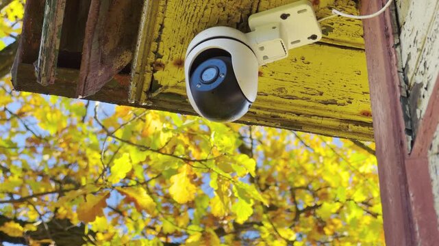A security camera hangs under a weathered wooden roof facing yellow autumn leaves. The steady surveillance creates a reassuring sense of safety and calm order.