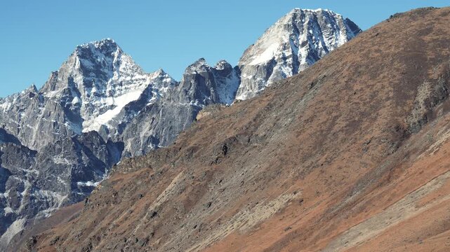 Majestic snow capped peaks of himalayas revealing themselves from behind barren, reddish-brown mountain ridge under clear blue sky, capturing grandeur of the alpine landscape