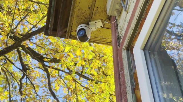 A white surveillance camera mounts beneath an old wooden roof near golden autumn trees. Its quiet presence brings a comforting feeling of security and peaceful safety.