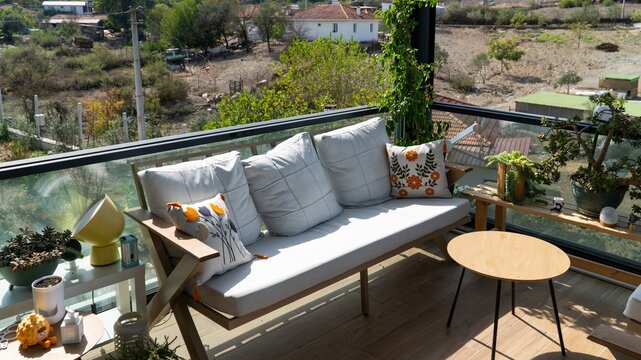 Cozy terrace with a wooden sofa and cushions, embroidered pillows, potted plants and a round coffee table, overlooking a green village on a sunny day