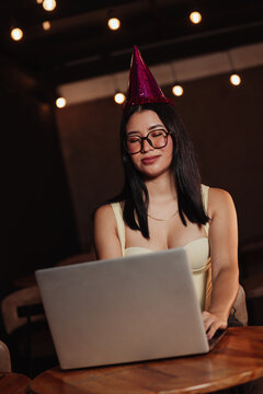 Beautiful woman wearing a birthday hat, using her laptop in a bar. Concept: working on one's birthday.