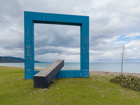 Aerial view of Monumento per un Poeta Morto, a large blue square sculpture framing the sea, set on a grassy coastline with a tall agave plant in Villa Margi, Sicilia, Italy.