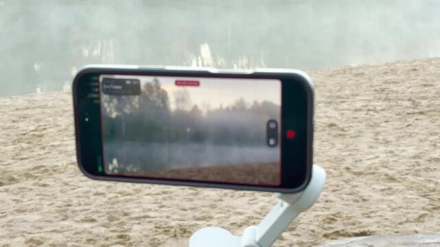 A smartphone captures a misty park lake on a sandy beach in Pushkin, Russia, in slow motion. The quiet foggy view creates a calm, tranquil mood.