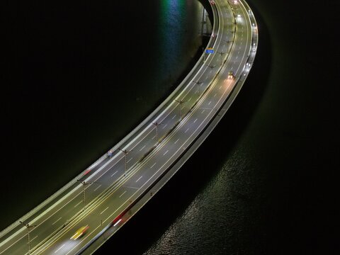 Aerial view of Bali Mandara Toll Road at night with illuminated street lamps and light trails from moving vehicles over the dark water in Kecamatan Kuta Selatan, Bali, Indonesia.