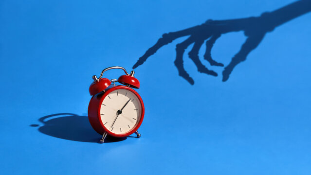 Twin bell alarm clock on blue, skeleton finger shadow about to touch it. Time running out, deadline pressure