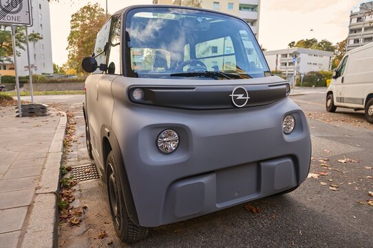 Small grey matte Opel Rocks Electric microcar parked on a city street sidewalk during autumn day with modern apartment buildings background.