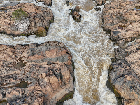 Aerial view of the turbulent Li Phi Somphamit Waterfalls with rushing brown water flowing through jagged rock formations and small islands in Khon Tai, Champasak Province, Laos.