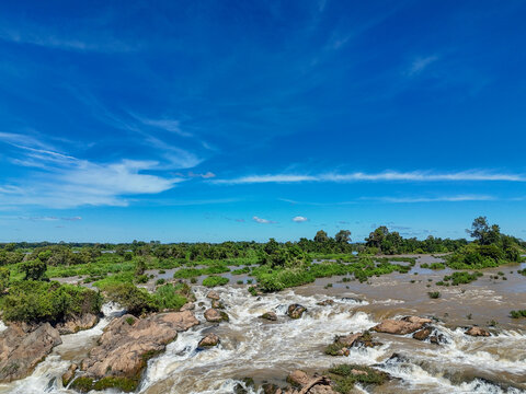 Aerial view of Li Phi Somphamit Waterfalls with rushing river water cascading over rocky outcrops surrounded by lush green vegetation under a blue sky in Khon Tai, Champasak Province, Laos.