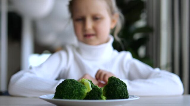 Young girl hesitates over plate of broccoli at table, making displeased face before reaching toward dinner. Child shows reluctance to eat vegetables, familiar parenting challenge at mealtime.