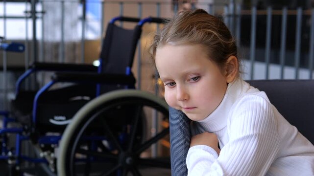 Young girl sits beside wheelchair, resting her head on arm with subdued expression. Child with disability awareness theme leans quietly in bright institutional room with mobility equipment nearby.