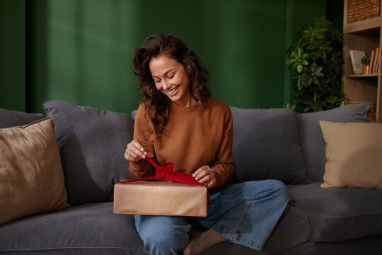 Happy young woman sitting on a gray sofa unties a red bow on a gift.