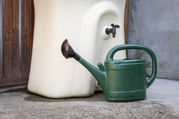 Green watering can stands beside a cream rainwater barrel with a tap against a rustic wooden door. A water saving concept promotes rainwater harvesting and sustainable gardening at home. © FrenchDailyLife