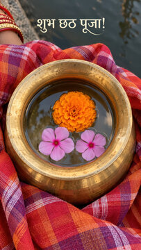 brass kalash pot filled with clear water holding a bright orange marigold blossom and two pink periwinkle flowers resting on traditional red orange gamcha cloth with Shubh Chhath Puja text in Hindi