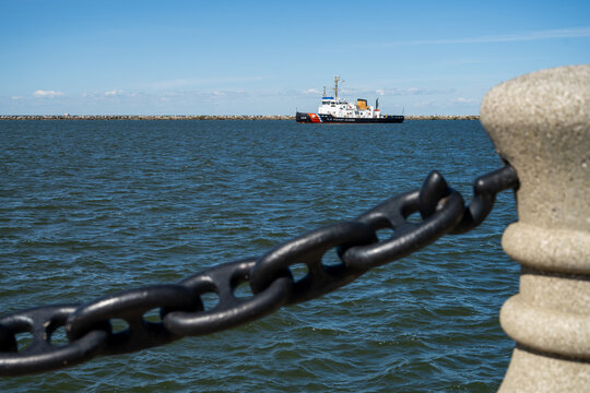 Cleveland, United States - 08 October 2026: View of a US Coast Guard cutter patrolling Lake Erie near the breakwater, framed by a heavy black metal chain and concrete bollard.