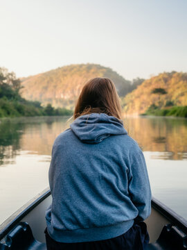 Kecamatan Kambera, Indonesia - 02 August 2026: View of a person in a grey hoodie sitting in a small boat on a calm river with rolling hills in the background.