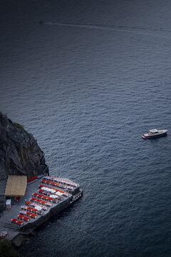 Amalfi, Campania, Italy - 14 October 2026: View of a cliffside beach club terrace with rows of red sun loungers and umbrellas overlooking the blue Tyrrhenian Sea, with a small boat nearby.