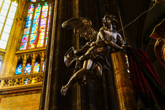 Prague, Czechia - 05 October 2026: View of a detailed metal angel statue suspended inside St. Vitus Cathedral, with vibrant stained glass windows casting colorful light in the background.