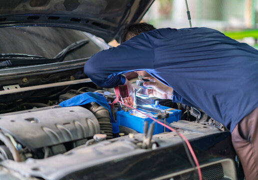 Auto mechanic using a headlamp flashlight to inspect and repair a blue car battery under an open hood.