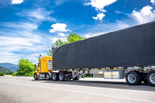 Bright yellow low cab profile big rig semi truck transporting cargo in conestoga flat bad semi trailer running on the flat highway road