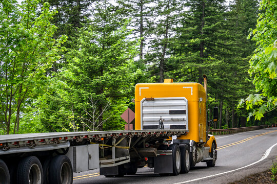 Classic yellow big rig semi truck with empty flat bed semi trailer running on the winding mountain road with green forest