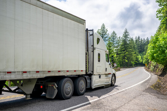 Commercial carrier white big rig semi truck transporting cargo in dry van semi trailer driving on the winding narrow road at sunny day