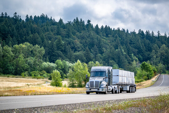 Dark gray bonnet brig semi truck transporting fastened covered cargo on the long flat bed semi trailer running on the narrow highway road with forest on the hill