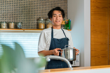 キッチンで鍋を持ち笑顔を見せるエプロン姿の男性。料理や家事を楽しむ暮らしのイメージ © buritora