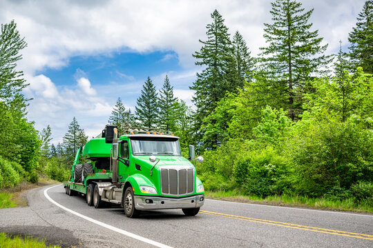Green day cab big rig semi truck transporting bulky tractor on heavy duty step deck semi trailer carefully running on winding narrow highway road
