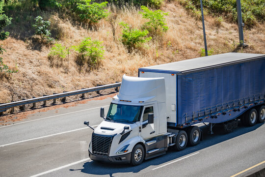 Innovative high-tech white big rig day cab semi truck transporting cargo in conestoga flat bed semi trailer running on the highway road intersection