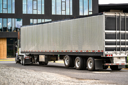 Powerful classic gray big rig semi truck transporting cargo in corrugated refrigerator semi trailer running on the urban city street with multilevel apartment buildings