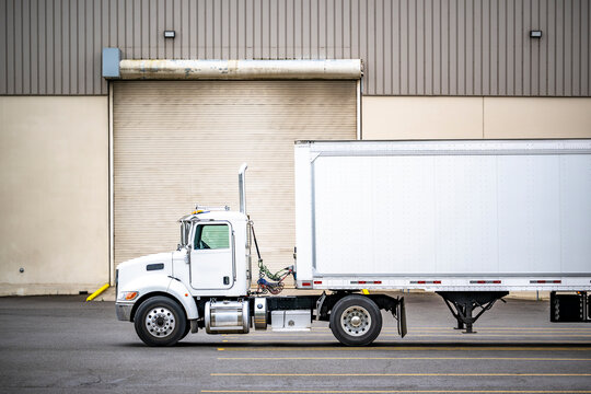 Profile of the clean day cab big rig semi truck with dry van semi trailer standing on the warehouse parking lot waiting for the next load