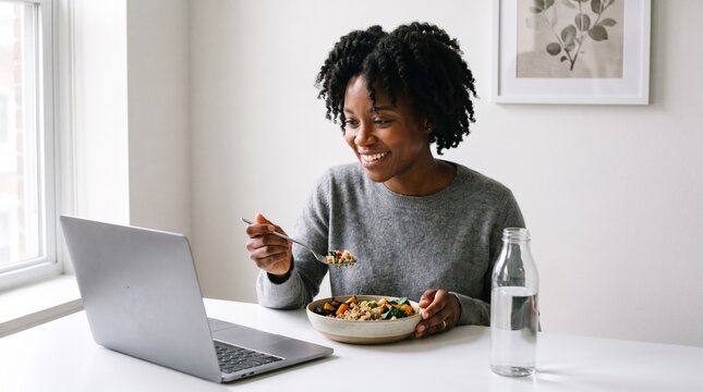 Black woman eating healthy lunch watching laptop video, working remotely from home, bright modern interior living room office