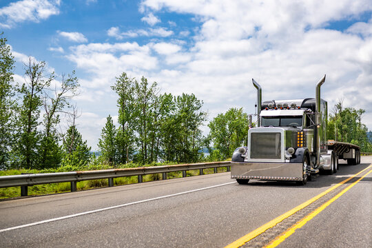 Style icon black big rig semi truck tractor with huge chrome exhaust pipes and light added accents transporting empty flat bed semi trailer running on the straight divided highway road