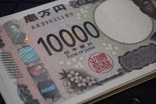 A close-up shot focusing on a Japanese 10000 yen currency banknote showing serial numbers, the Bank of Japan text, a red official seal, and a holographic foil security feature.