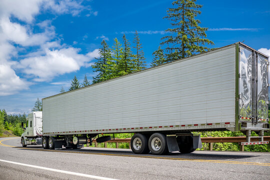 White low cab profile big rig commercial semi truck transporting cargo in long refrigerator semi trailer moving on the winding road at sunny day
