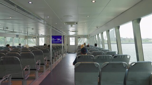 Passenger ferry cabin in New York City, New York, USA. Rows of gray seats, a few commuters, big windows over the East River and an industrial shore with smokestacks. Public water transit on a hazy day