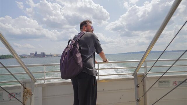 Bearded man with purple backpack leans on ferry stern rail in New York City, New York, USA. White wake, harbor water and distant waterfront buildings. Sunny summer day, fluffy clouds, travel.