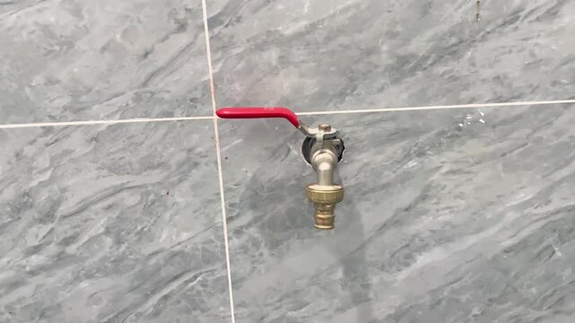 A metallic water tap faucet with a red lever handle mounted on a grey marbled tile wall. A close-up shot of functional plumbing hardware installed on a glossy bathroom or outdoor kitchen wall backdrop
