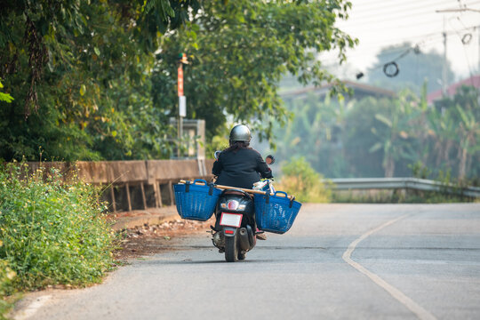 Unrecognizable local Thai farmer in helmet driving on motorcycle carrying plastic baskets with harvest, man worker riding along countryside road on scooter with containers of produce in Thailand.